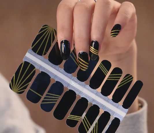 Black nail wraps with gold patterns on a hand against a neutral background